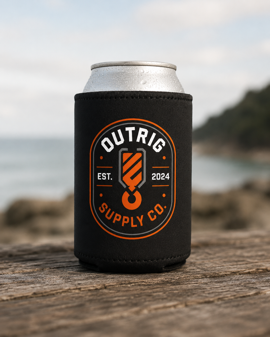 Black Outrig Supply Co stubby cooler with orange-and-white badge on a can at the beach.