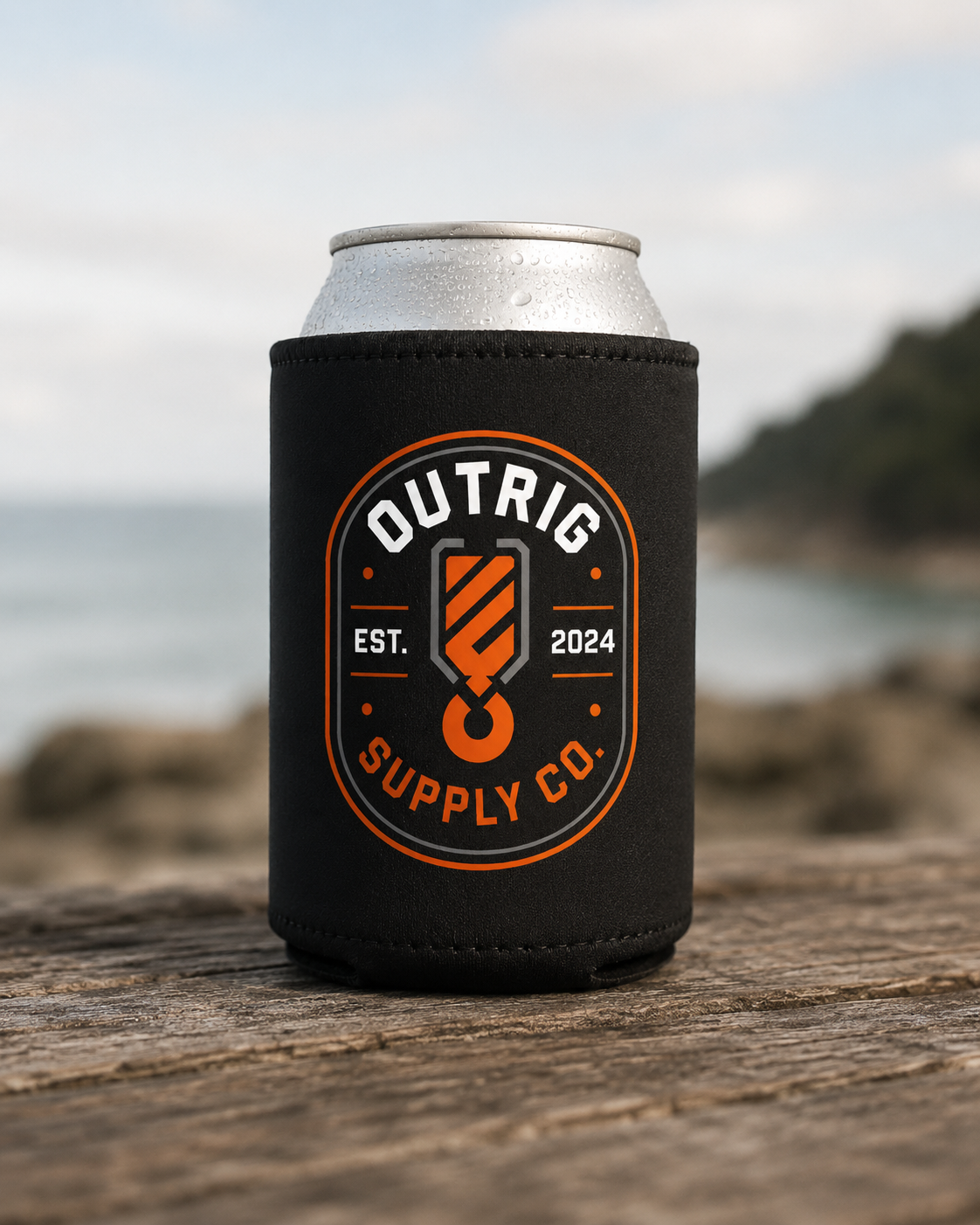 Black Outrig Supply Co stubby cooler with orange-and-white badge on a can at the beach.
