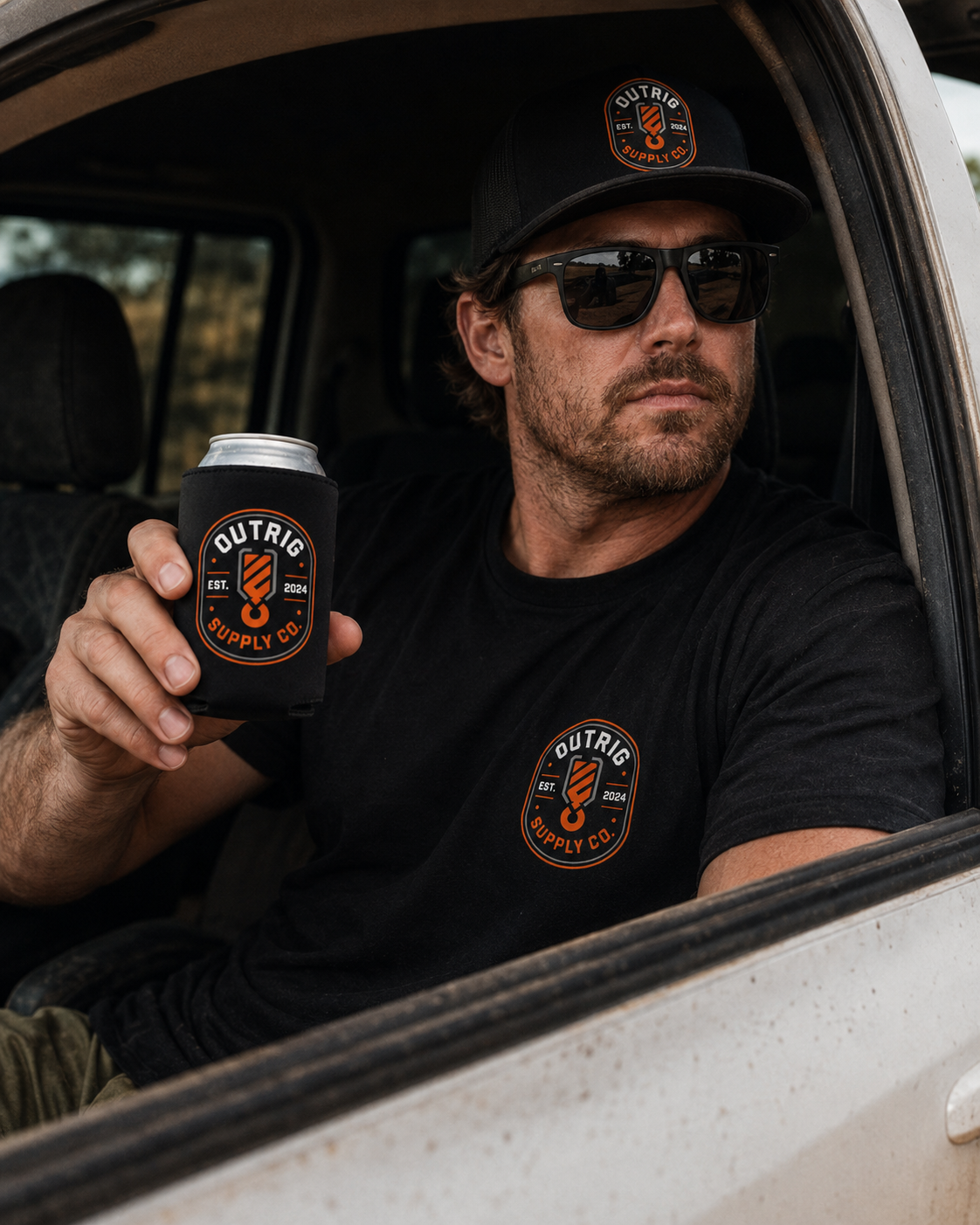 Man wearing sunglasses and an Outrig cap holding a black Outrig Supply Co stubby cooler inside a vehicle.